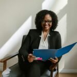Professional African American woman with glasses and curly hair holding a blue folder indoors.